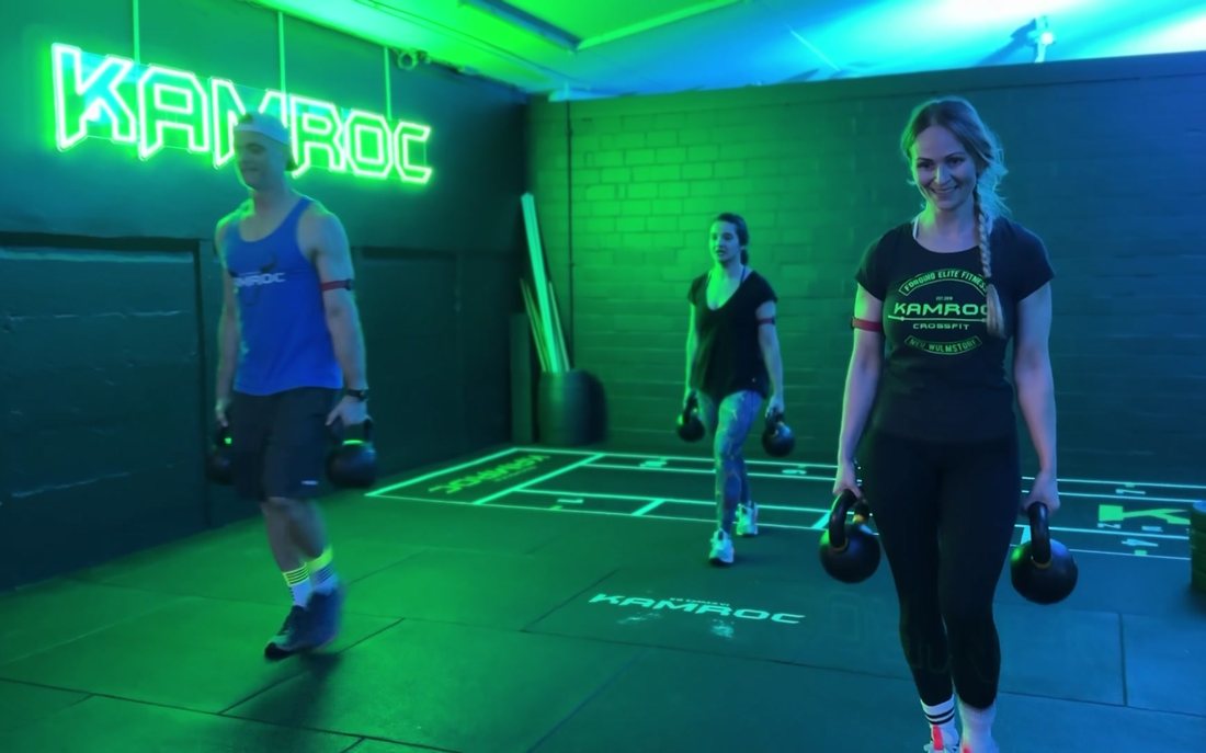 Farmers Carry im Team bei KAMROC: drei Mitglieder tragen Kettlebells unterm Neon-Logo